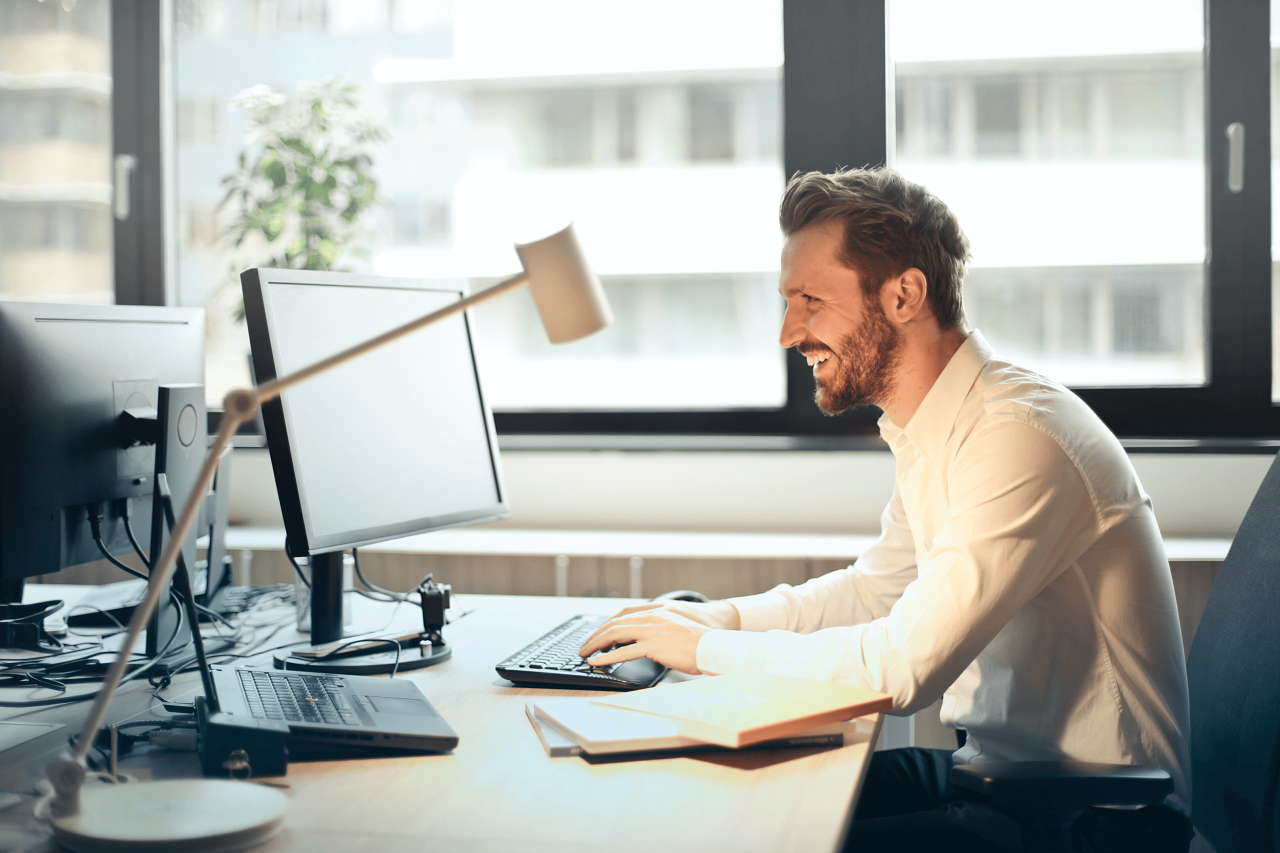 An excited man in a white shirt is sitting in his office and looking at his computer monitor.