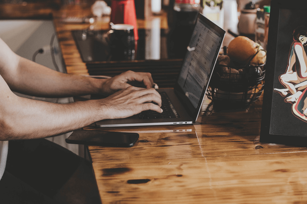 A person typing on a laptop. The laptop is on a wooden table. Next to it lies a mobile phone. 